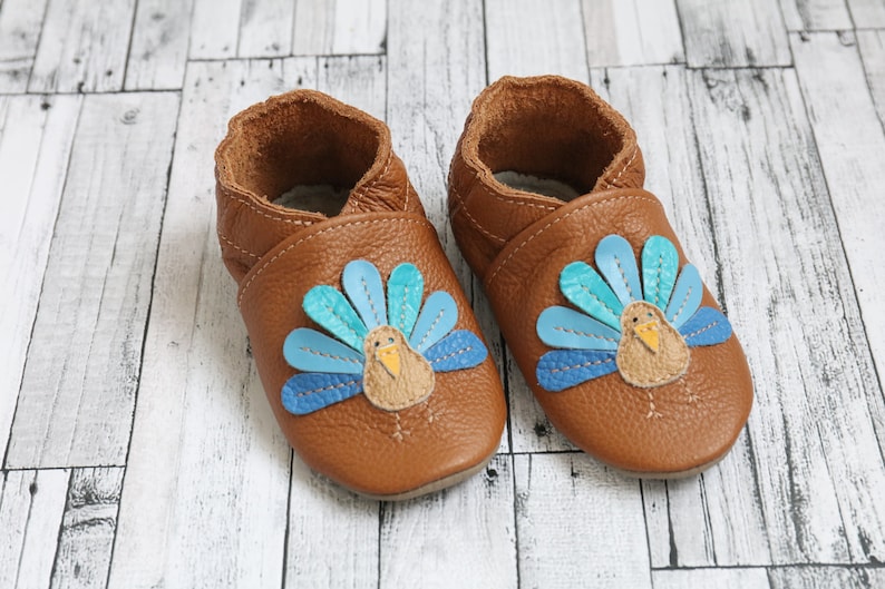 Puede incluir: Un par de zapatos de beb&eacute; de cuero marr&oacute;n con un dise&ntilde;o de pavo. El pavo tiene un dise&ntilde;o de plumas azules, verde azulado y amarillas.