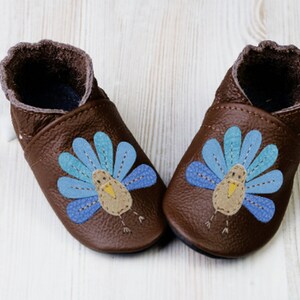 Puede incluir: Un par de zapatos de beb&eacute; de cuero marr&oacute;n con un dise&ntilde;o de pavo azul y amarillo.