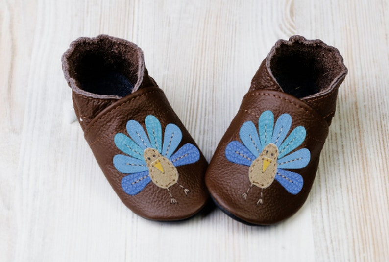 Puede incluir: Un par de zapatos de beb&eacute; de cuero marr&oacute;n con un dise&ntilde;o de pavo azul y amarillo.