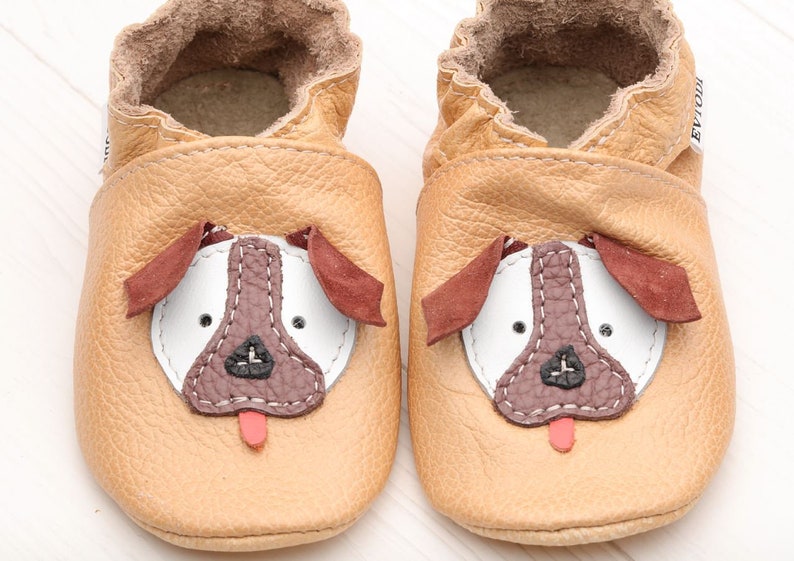 May include: A pair of tan leather baby shoes with a brown dog face design. The shoes have a white nose, brown eyes, and a red tongue.