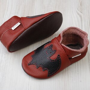 May include: A pair of red leather baby shoes with a black bat applique. The shoes have a soft sole and a dark brown interior. The brand name "EVTODI" is visible on a small tag.