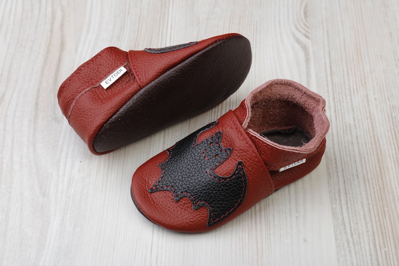 May include: A pair of red leather baby shoes with a black bat applique. The shoes have a soft sole and a dark brown interior. The brand name "EVTODI" is visible on a small tag.