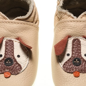 May include: A pair of beige leather baby shoes with a dog face design. The dog face features brown ears, a brown nose, and black eyes.