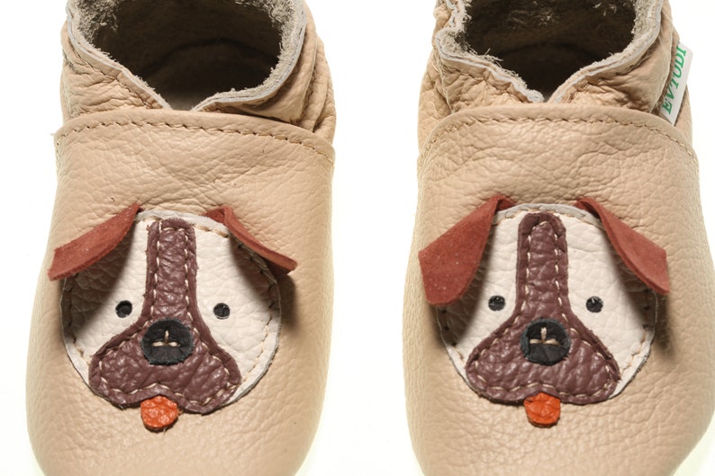 May include: A pair of beige leather baby shoes with a dog face design. The dog face features brown ears, a brown nose, and black eyes.