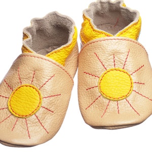 May include: A pair of baby moccasins with a sun design. The moccasins are made of beige leather with yellow sun accents and red stitching.