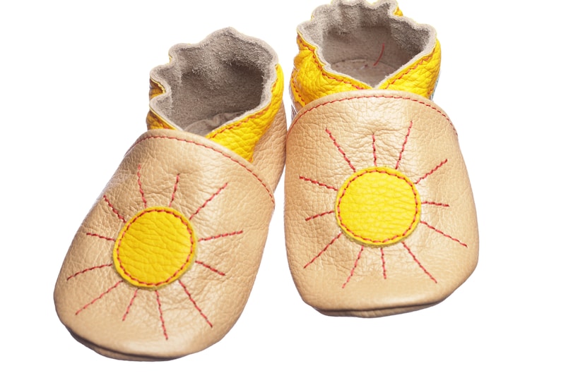 May include: A pair of baby moccasins with a sun design. The moccasins are made of beige leather with yellow sun accents and red stitching.