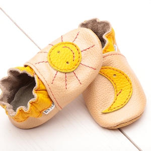 May include: A pair of baby shoes with a sun and moon design. The shoes are made of soft leather and have a yellow and beige color scheme.