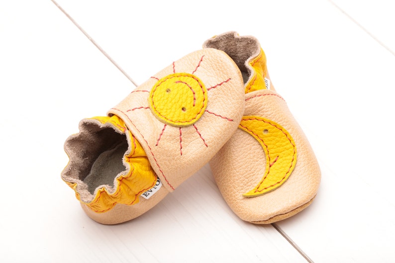 May include: A pair of baby shoes with a sun and moon design. The shoes are made of soft leather and have a yellow and beige color scheme.