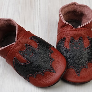 May include: A pair of burgundy leather baby shoes with a black bat applique on the toe. The bat is stitched with red thread. The shoes have a soft, flexible sole and a rolled cuff.