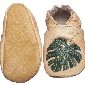 May include: A pair of tan leather baby shoes featuring a green monstera leaf design. The shoes have a soft, flexible sole and are ideal for little feet.