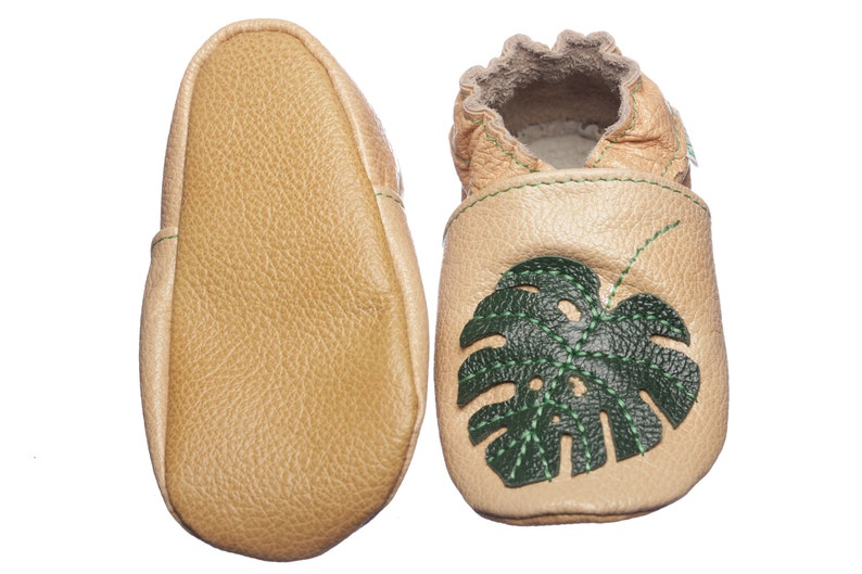 May include: A pair of tan leather baby shoes featuring a green monstera leaf design. The shoes have a soft, flexible sole and are ideal for little feet.