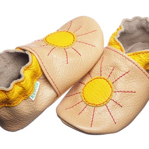 May include: A pair of beige leather baby shoes with yellow sun designs and red stitching. The shoes have a yellow trim around the top and a green label that says "Evtodi".