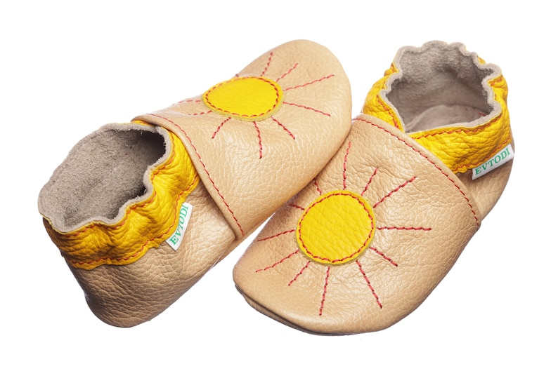 May include: A pair of beige leather baby shoes with yellow sun designs and red stitching. The shoes have a yellow trim around the top and a green label that says "Evtodi".