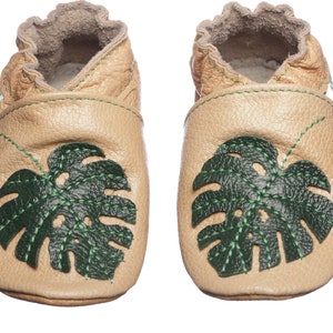 May include: A pair of tan leather baby shoes with green monstera leaf designs. The shoes have a soft, flexible sole and are perfect for crawling and walking.