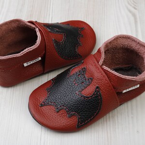 May include: A pair of burgundy leather baby shoes with a black bat applique stitched on the front. The shoes have a soft, flexible sole and a rolled cuff. The brand name "EVTODI" is printed on a small tag.