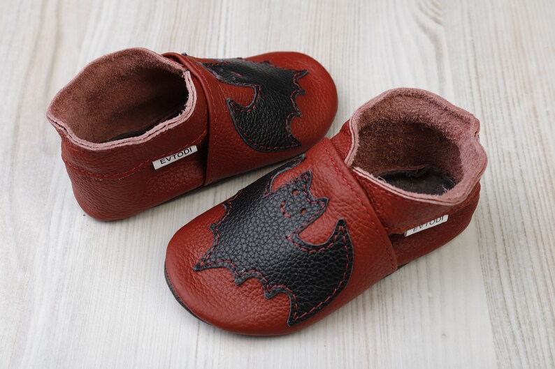 May include: A pair of burgundy leather baby shoes with a black bat applique stitched on the front. The shoes have a soft, flexible sole and a rolled cuff. The brand name "EVTODI" is printed on a small tag.