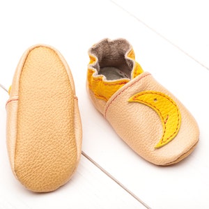 May include: A pair of baby shoes made of soft leather in a light beige color. One shoe has a yellow crescent moon design on the toe.