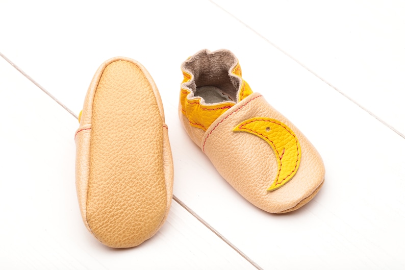 May include: A pair of baby shoes made of soft leather in a light beige color. One shoe has a yellow crescent moon design on the toe.