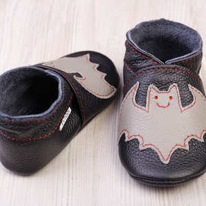 May include: A pair of black leather baby shoes with a gray bat applique stitched with red thread. The shoes have a soft, gray interior and are designed for infants. The soles are flexible.