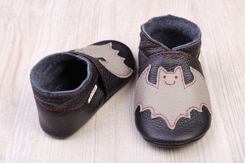May include: A pair of black leather baby shoes with a gray bat applique stitched with red thread. The shoes have a soft, gray interior and are designed for infants. The soles are flexible.