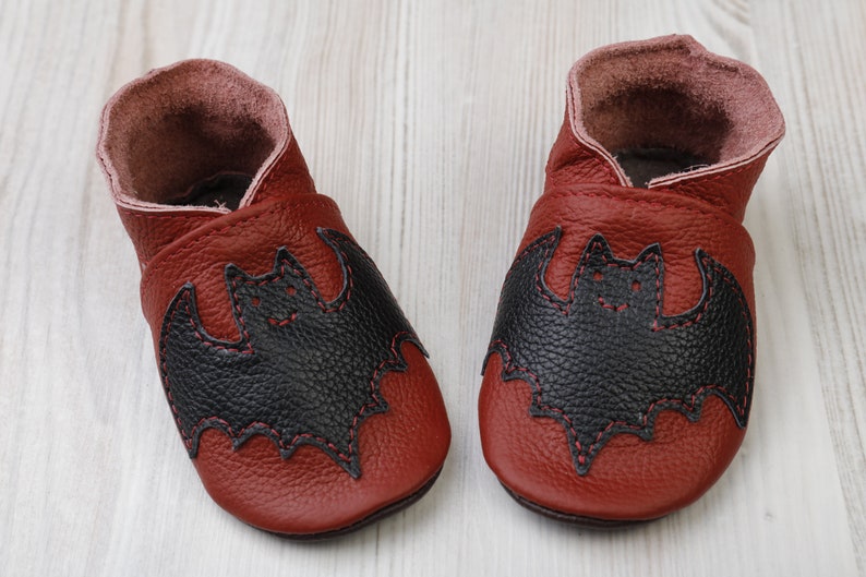 May include: A pair of red leather baby shoes with a black bat applique stitched on the front. The bat design is outlined with red thread. The interior of the shoes is a lighter shade of red.