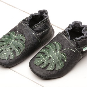 May include: Black leather baby shoes featuring a green monstera leaf design. The shoes have a soft, flexible sole, ideal for crawling and walking.