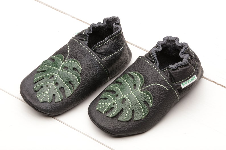 May include: Black leather baby shoes featuring a green monstera leaf design. The shoes have a soft, flexible sole, ideal for crawling and walking.