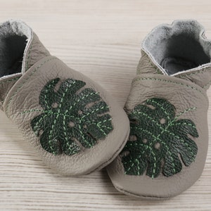 May include: A pair of grey leather baby shoes with green monstera leaf embroidery. The shoes have a soft, flexible sole and are perfect for little feet.