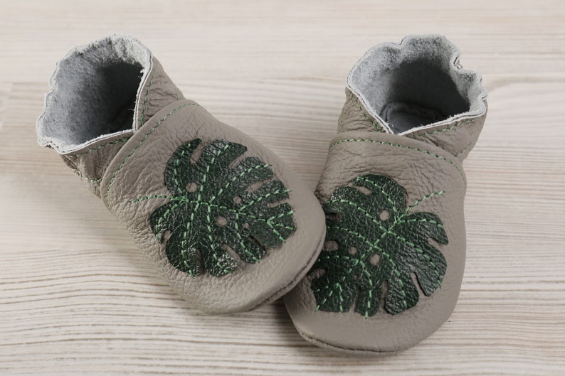 May include: A pair of grey leather baby shoes with green monstera leaf embroidery. The shoes have a soft, flexible sole and are perfect for little feet.