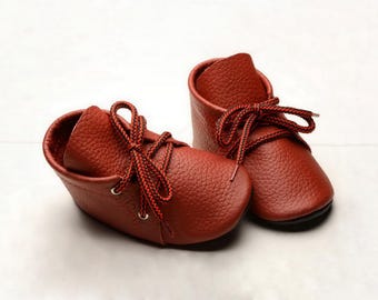May include: A pair of rust-colored leather shoes with a pebbled texture. The shoes feature high-top ankle coverage, rounded toes, and are secured with black and red patterned laces threaded through silver-toned metal eyelets on each side.