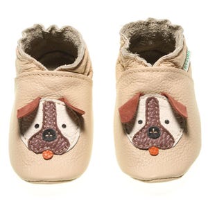 May include: A pair of beige leather baby shoes with a brown dog face design. The shoes have a soft, flexible sole and are perfect for first steps.