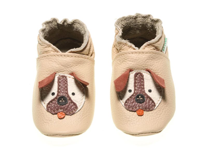 May include: A pair of beige leather baby shoes with a brown dog face design. The shoes have a soft, flexible sole and are perfect for first steps.