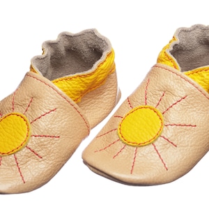 May include: A pair of beige leather baby shoes with a yellow sun design and red stitching. The shoes have a yellow trim around the top and the brand name "EVTODI" is printed on the side.