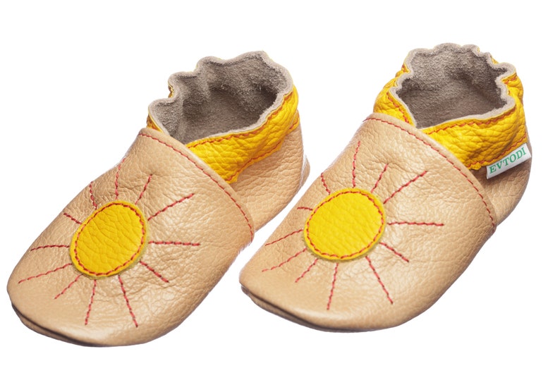 May include: A pair of beige leather baby shoes with a yellow sun design and red stitching. The shoes have a yellow trim around the top and the brand name "EVTODI" is printed on the side.