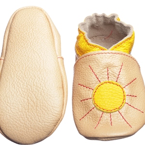 May include: A pair of baby shoes with a sun design. The shoes are made of soft leather and have a yellow sun with red stitching on the front. The soles are beige.
