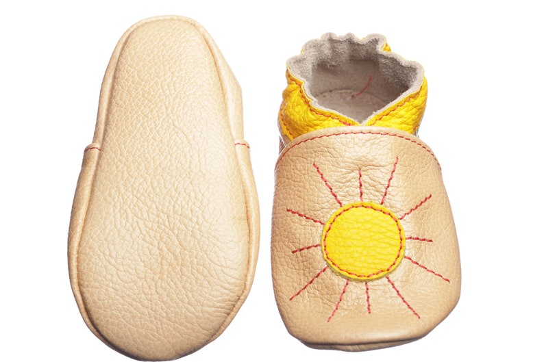 May include: A pair of baby shoes with a sun design. The shoes are made of soft leather and have a yellow sun with red stitching on the front. The soles are beige.