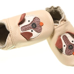 May include: A pair of beige leather baby shoes with a dog face design. The shoes have brown and white accents and a small orange button nose.