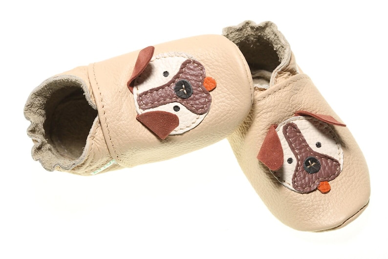 May include: A pair of beige leather baby shoes with a dog face design. The shoes have brown and white accents and a small orange button nose.