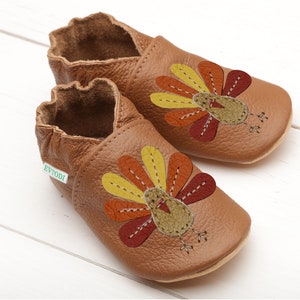 Puede incluir: Un par de zapatos de beb&eacute; de cuero marr&oacute;n con un dise&ntilde;o de pavo. El pavo est&aacute; bordado con plumas amarillas, naranjas y rojas. Los zapatos tienen una etiqueta verde que dice "Evyodi".
