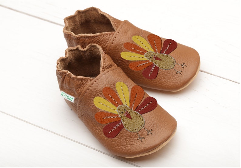 Puede incluir: Un par de zapatos de beb&eacute; de cuero marr&oacute;n con un dise&ntilde;o de pavo. El pavo est&aacute; bordado con plumas amarillas, naranjas y rojas. Los zapatos tienen una etiqueta verde que dice "Evyodi".