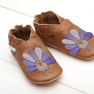 Puede incluir: Un par de zapatos de beb&eacute; de cuero marr&oacute;n con un dise&ntilde;o de pavo morado y gris. Los zapatos tienen una etiqueta verde que dice "Evodi".