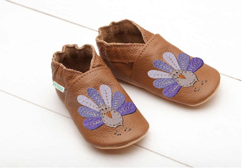 Puede incluir: Un par de zapatos de beb&eacute; de cuero marr&oacute;n con un dise&ntilde;o de pavo morado y gris. Los zapatos tienen una etiqueta verde que dice "Evodi".