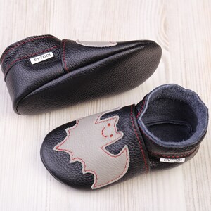 May include: Black leather baby shoes with a gray bat applique and red stitching. The shoes have a soft sole and a dark blue interior. The brand name "EVTODI" is visible on the side of the shoe.