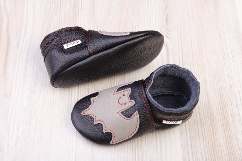 May include: Black leather baby shoes with a gray bat applique and red stitching. The shoes have a soft sole and a dark blue interior. The brand name "EVTODI" is visible on the side of the shoe.