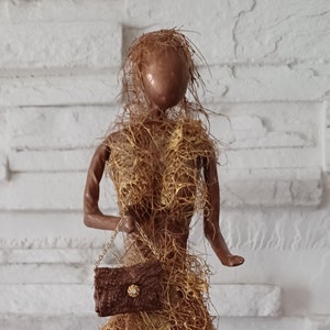 Peut inclure: Une figurine de femme brune et dorée portant une longue robe en fibres tissées. Elle tient un petit sac à main brun avec une chaîne dorée.
