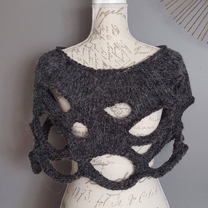 Könnte beinhalten: Ein grauer Strick-Schal mit einem einzigartigen Lochmuster. Der Schal wird auf einer Schaufensterpuppe mit einem Vintage-gedruckten Oberkörper präsentiert.