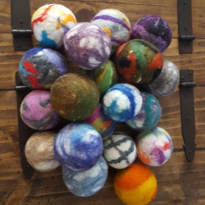 May include: A collection of colorful wool dryer balls, featuring various patterns and colors, including blue, green, purple, orange, and white. The balls are arranged in a pyramid shape on a wooden surface.