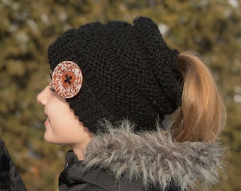 May include: A black knit beanie with a ponytail hole and a decorative wooden button. The beanie is worn by a person with long brown hair.