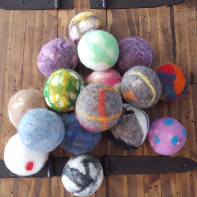May include: A collection of 15 colorful felt balls in various patterns and colors, including green, blue, pink, purple, gray, and brown. The balls are arranged in a pile on a wooden surface.