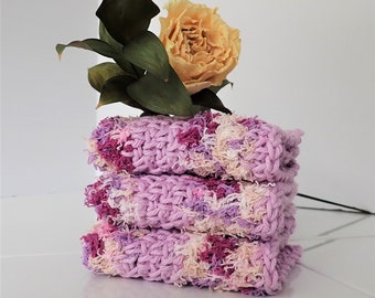 May include: A stack of three handmade washcloths in shades of lavender, pink, and cream. The washcloths are made of a textured, looped yarn. A dried yellow flower and green leaves are placed on top.